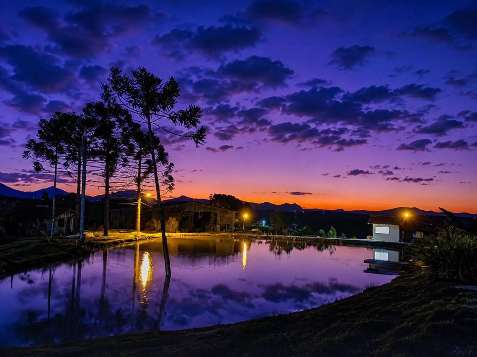Lago refletindo o céu roxo e laranja do anoitecer, com as luzes do sítio acesas