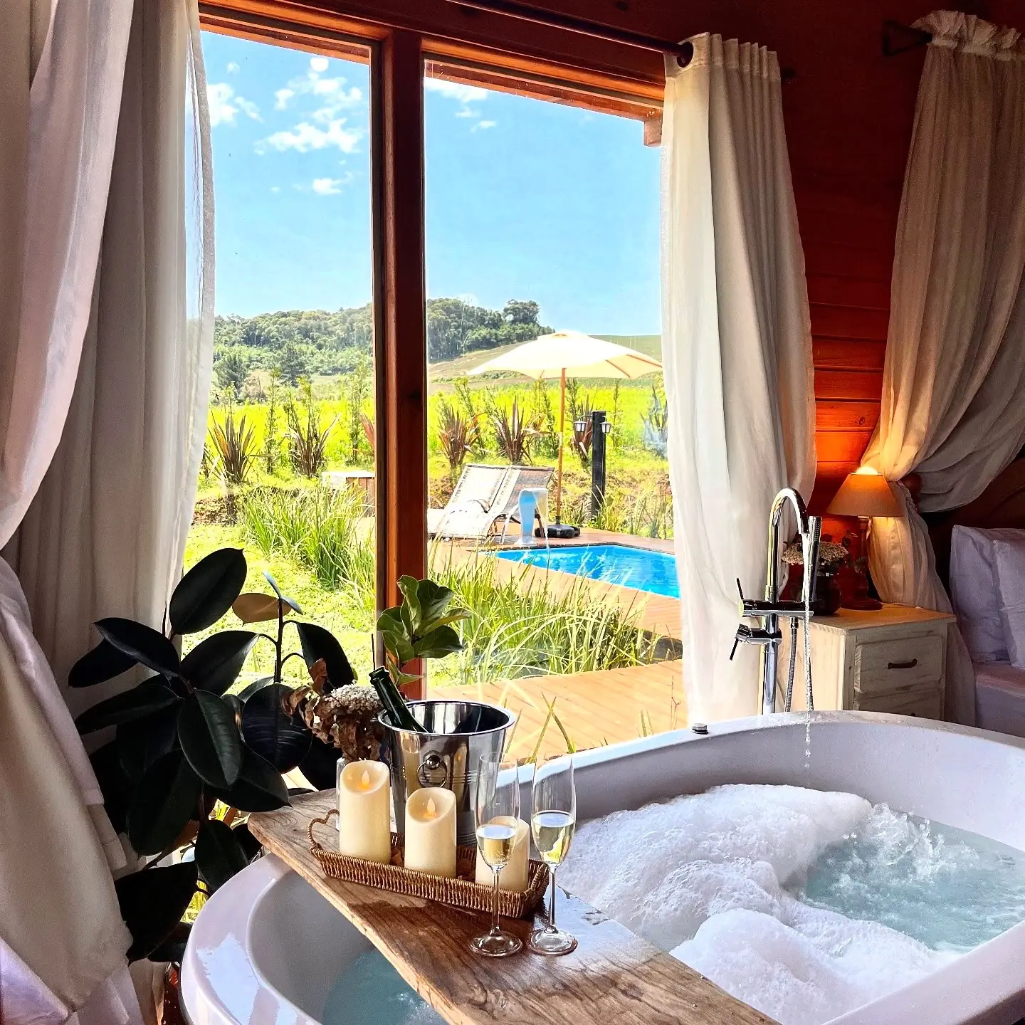 Banheira com espuma e espumante, de frente para a janela com vista para a piscina e o campo