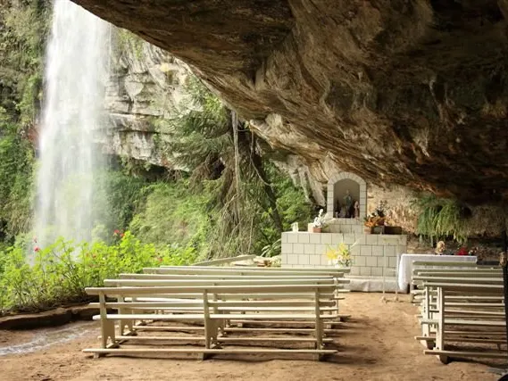 Cachoeira caindo ao lado da gruta, com os bancos da capela embaixo da pedra